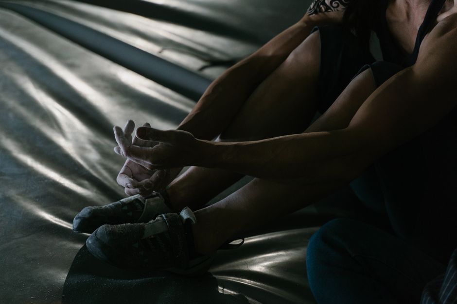 A close-up view of a seated individual wearing dark athletic shoes, with their legs extended and slightly bent at the knees. The person is dressed in dark clothing, and their hands are clasped togethe