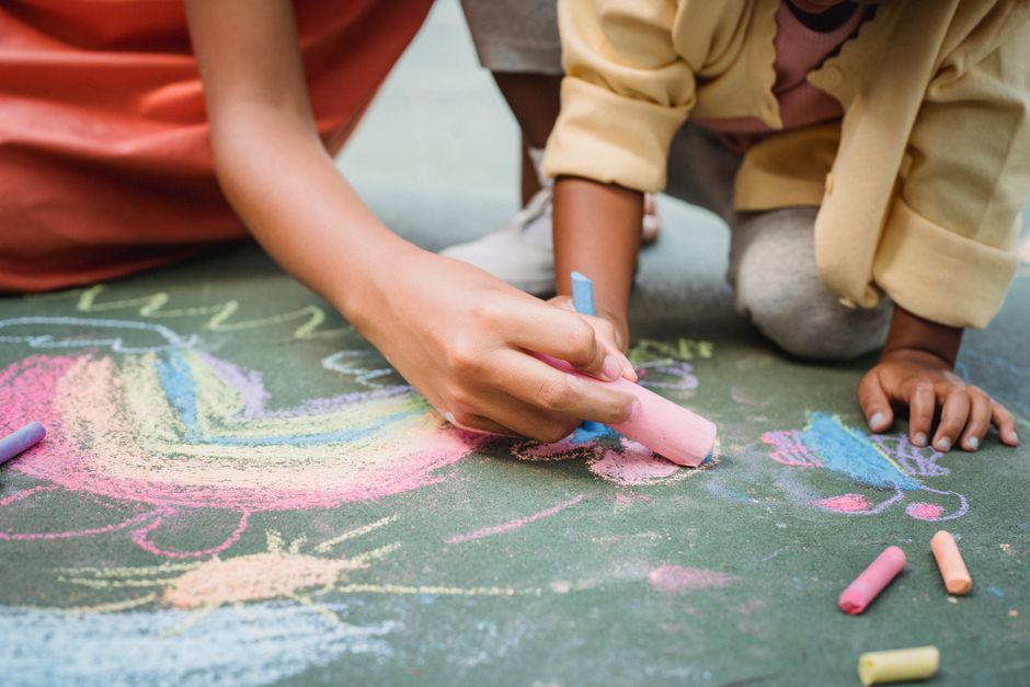 Close-up of two children drawing on a chalkboard with colorful chalks outdoors, with the focus on their hands and the vibrant chalk artwork. One child is holding a pink chalk, creating a colorful desi
